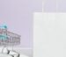 Minimalist image of a white shopping bag and mini cart on a soft pastel background.